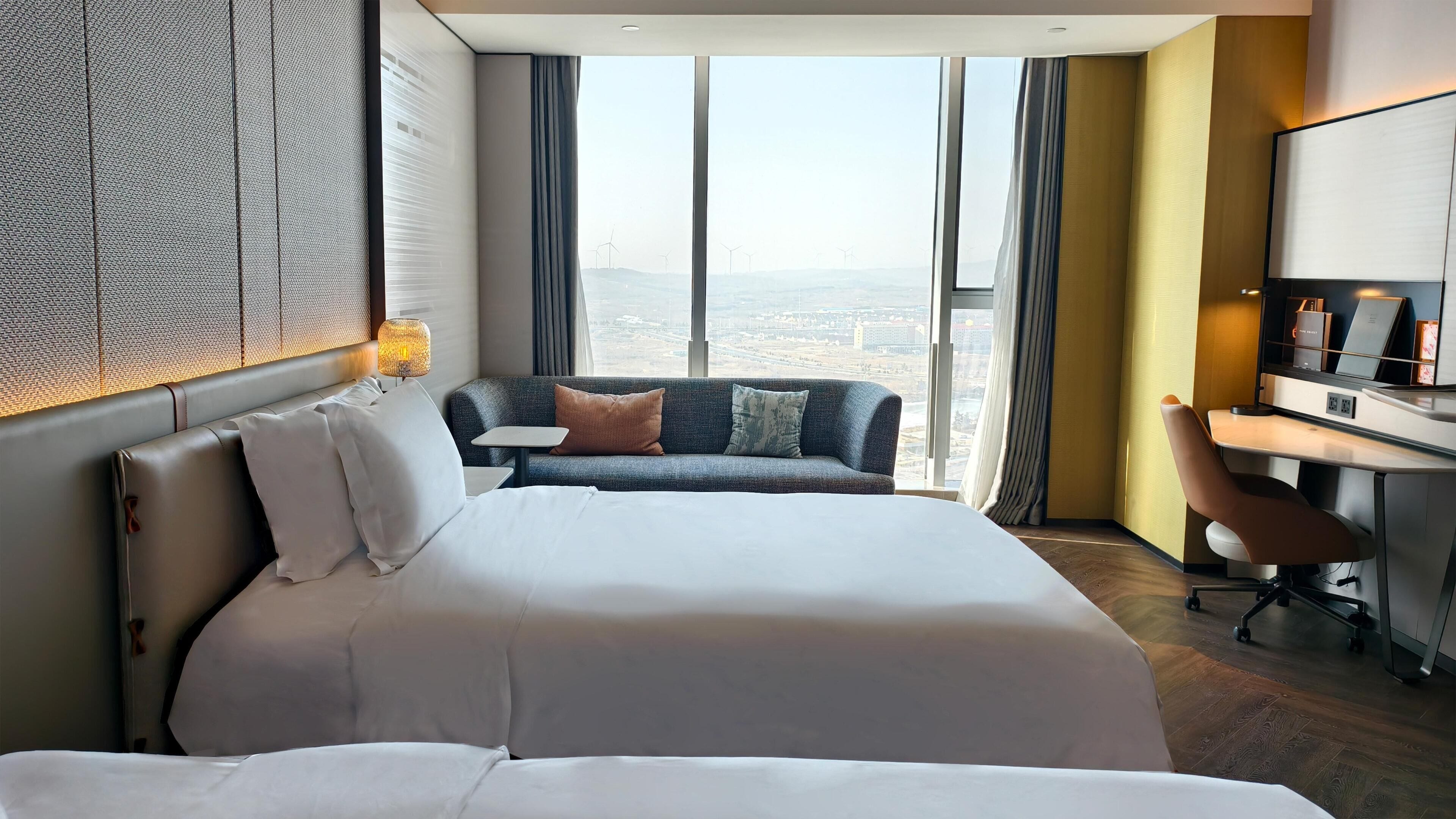 Habitación de hotel con una cama grande, un sofá, un escritorio y vistas a la ciudad a través de amplias ventanas.