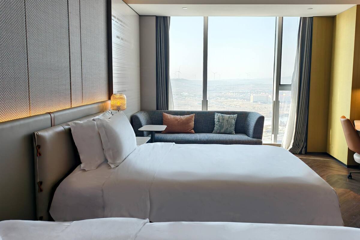 Une chambre d’hôtel avec un grand lit, un canapé, un bureau et une vue sur le paysage urbain grâce à de grandes fenêtres.