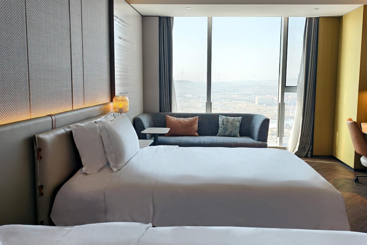 Habitación de hotel con una cama grande, un sofá, un escritorio y vistas a la ciudad a través de amplias ventanas.