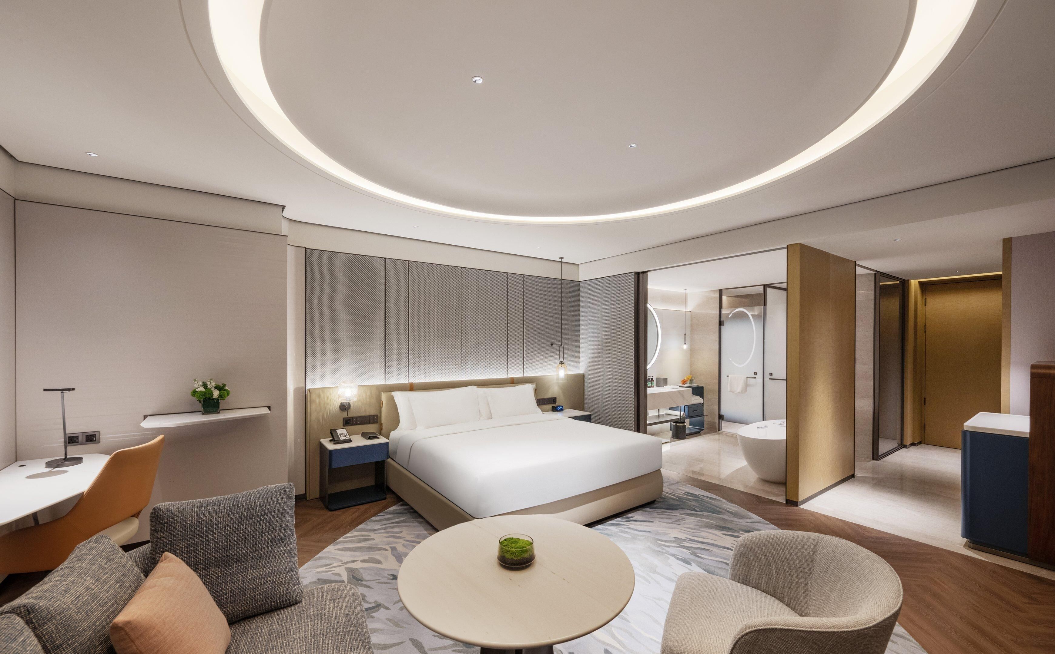 Una habitación de hotel moderna con cama, mesitas de noche, un escritorio, una silla, una mesa redonda y un baño con bañera e inodoro.