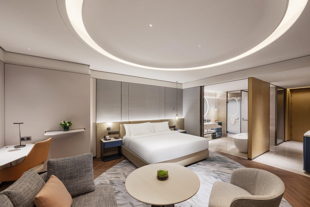 Une chambre d’hôtel moderne équipée d’un lit, de tables de chevet, d’un bureau, d’une chaise, d’une table ronde et d’une salle de bain avec baignoire et toilettes.