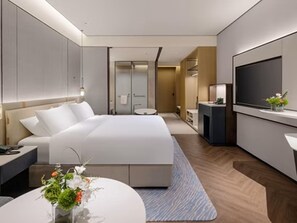 Et moderne hotelværelse med en stor seng, et skrivebord, et fjernsyn og et badeværelse, der kan ses igennem en glasdør.