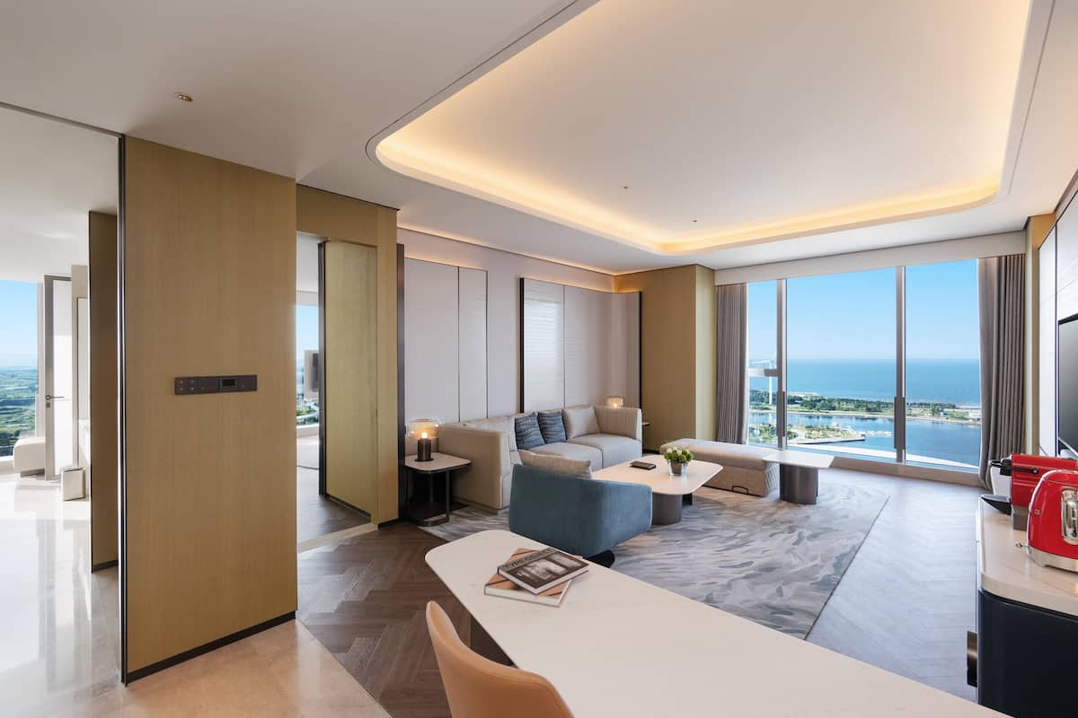 Une chambre d’hôtel moderne dotée d’un canapé, d’une table basse et d’un coin repas. Elle offre de grandes fenêtres avec vue sur l’océan et sur le paysage urbain.
