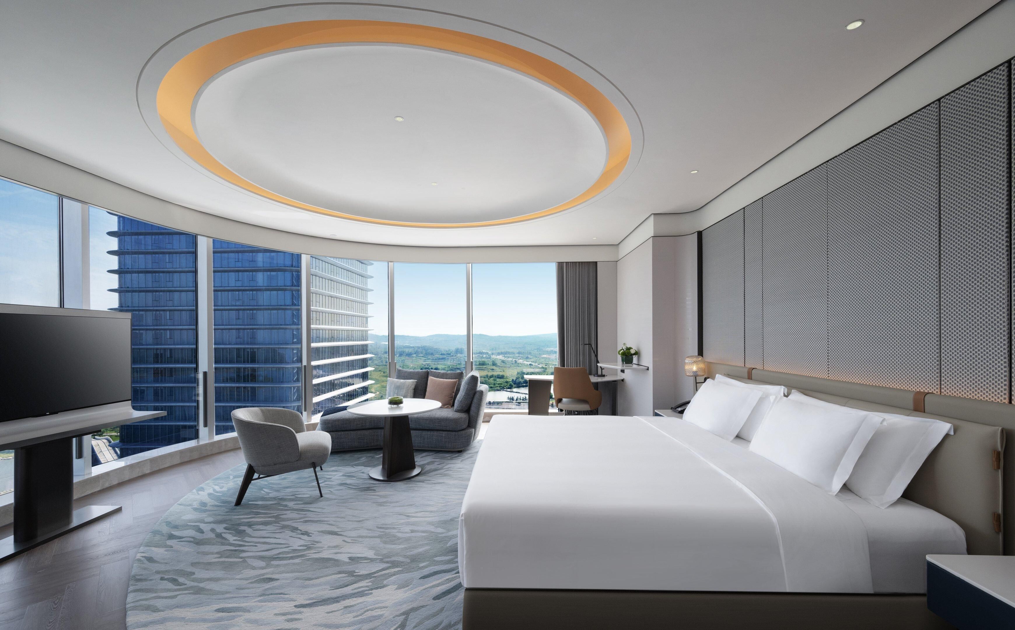 Una habitación de hotel moderna con una cama grande, un área de descanso con una mesa, un televisor y vistas a la ciudad.