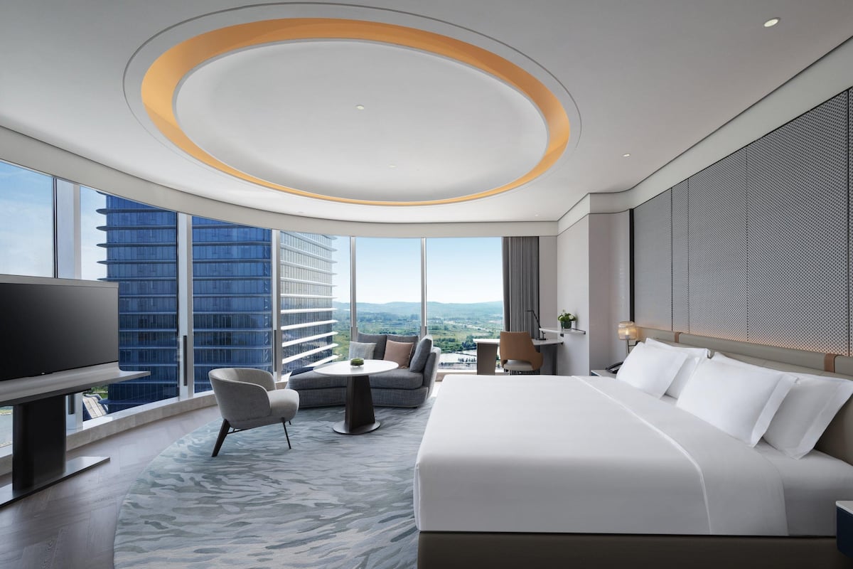 Una habitación de hotel moderna con una cama grande, un área de descanso con una mesa, un televisor y vistas a la ciudad.