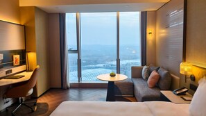 Et hotelværelse med et stort vindue, en seng, et skrivebord, en stol, en sofa og et lille bord med en plante.