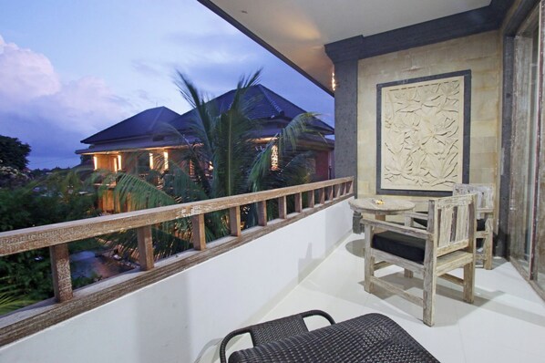 A balcony with a table and chairs, a decorative wall panel, and a view of a house at dusk. - Batu Empug Hotel (Ubud)