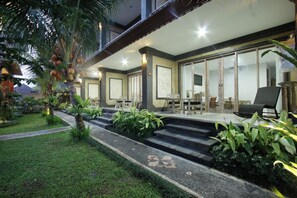 A well-lit outdoor area with steps leading to a covered seating area, surrounded by lush greenery and palm trees. - Batu Empug Hotel (Ubud)