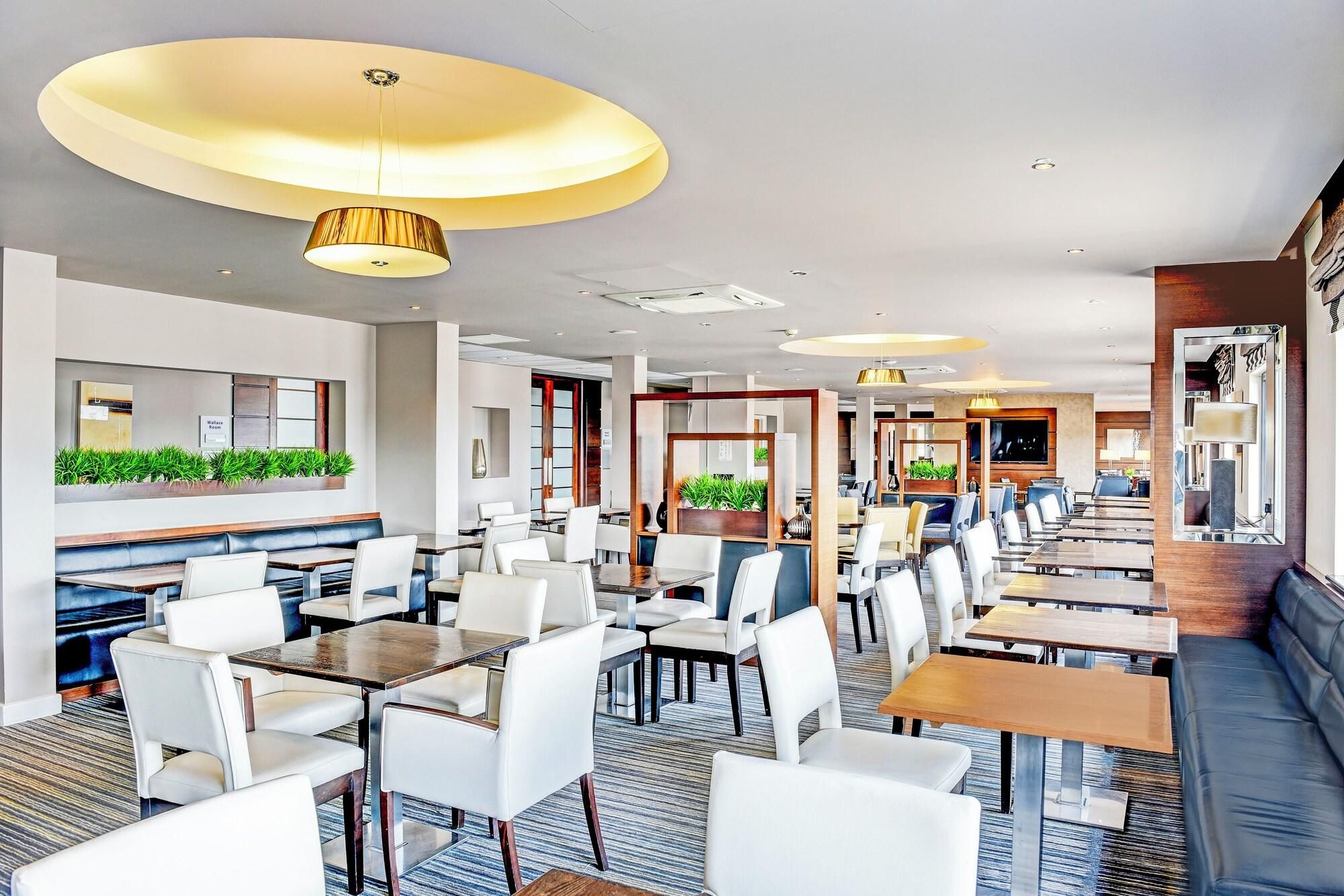Un hall d’hôtel moderne offrant différents types de sièges, notamment des tables et des banquettes, un espace d’accueil et un grand miroir.