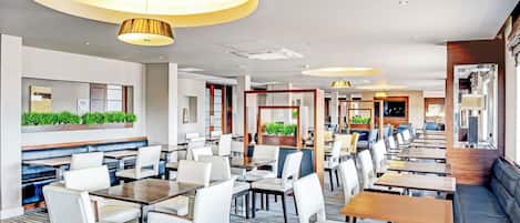 Un hall d’hôtel moderne offrant différents types de sièges, notamment des tables et des banquettes, un espace d’accueil et un grand miroir.