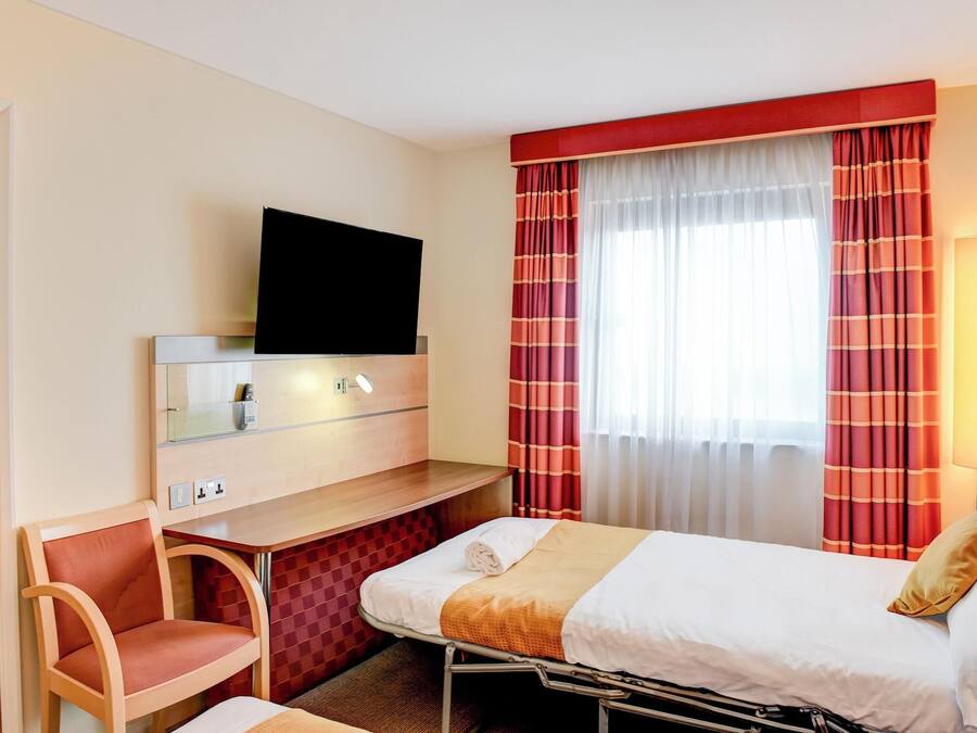 A hotel room with a bed, a desk with a flat-screen TV, a chair, and a window with striped curtains.