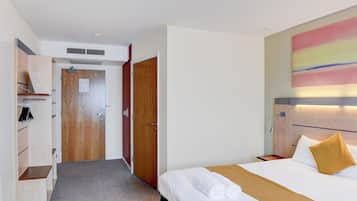 Una habitación de hotel con cama, mesita de noche, lámpara, una obra de arte en la pared y un pasillo que conduce a una puerta.