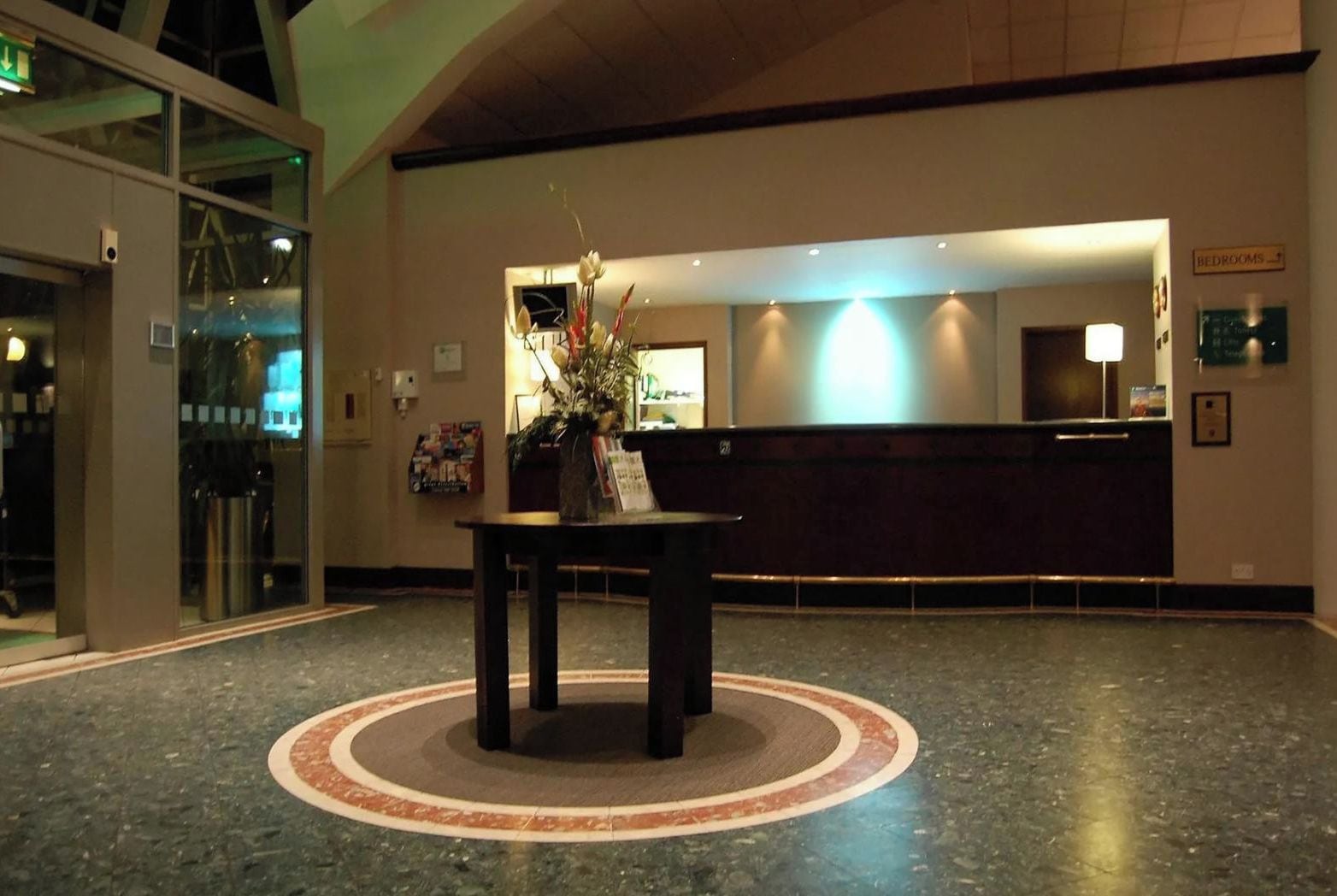 Un vestíbulo de hotel con un mostrador de recepción, un separador de vidrio y una mesa decorativa con flores.