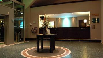 Un vestíbulo de hotel con un mostrador de recepción, un separador de vidrio y una mesa decorativa con flores.