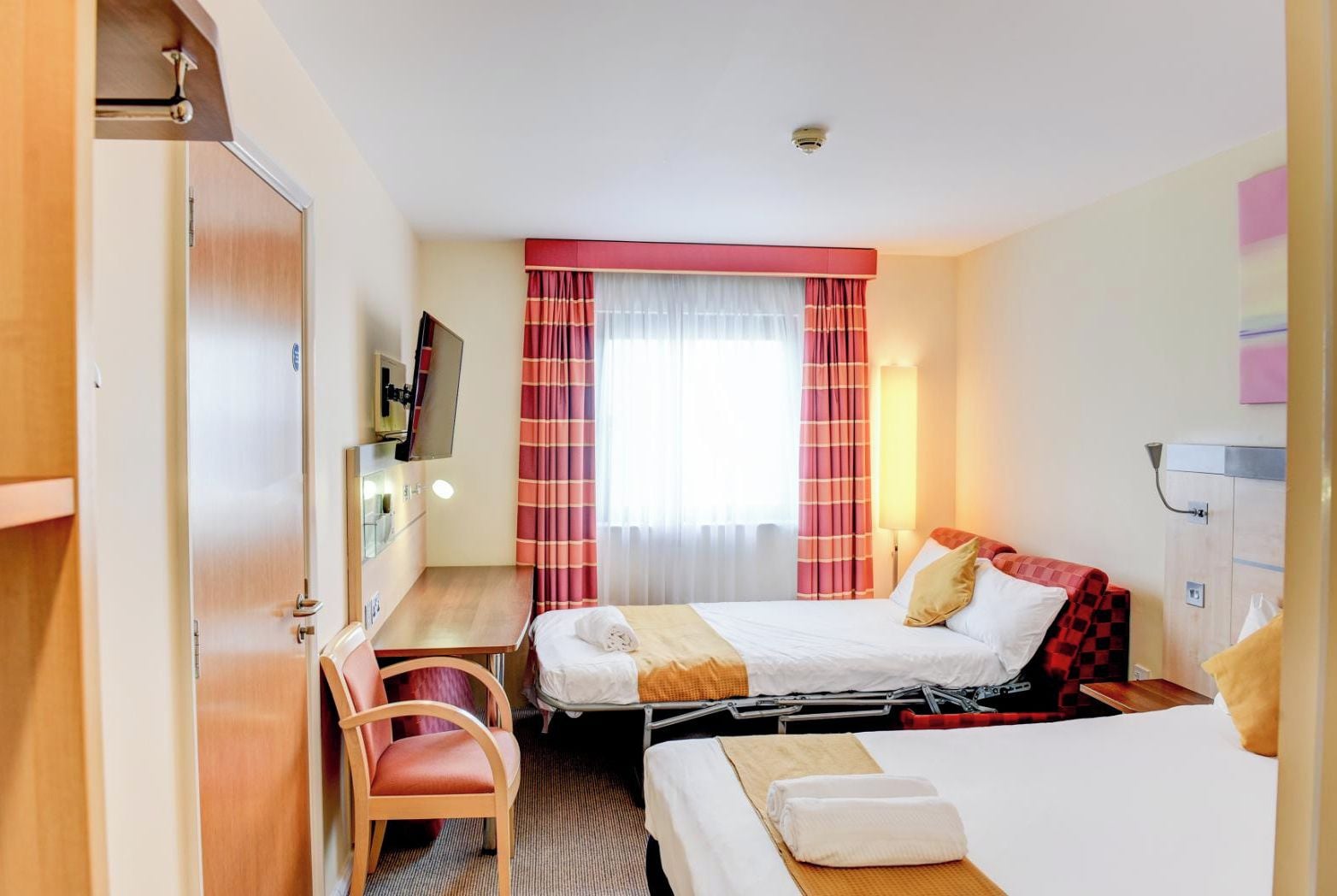A hotel room with two beds, a desk, a chair, a TV, and a window with curtains.