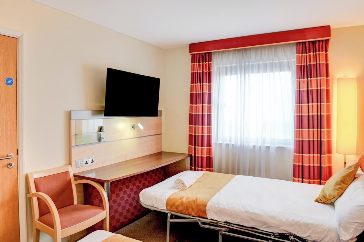 A hotel room with a bed, a desk, a chair, a TV, and a window with curtains.