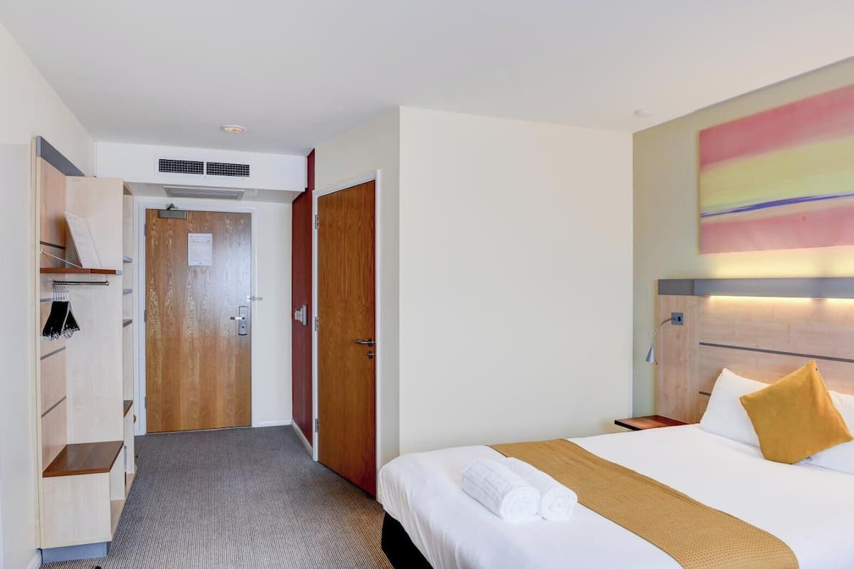 A hotel room with a bed, a headboard with a light fixture, a wardrobe, and a door.