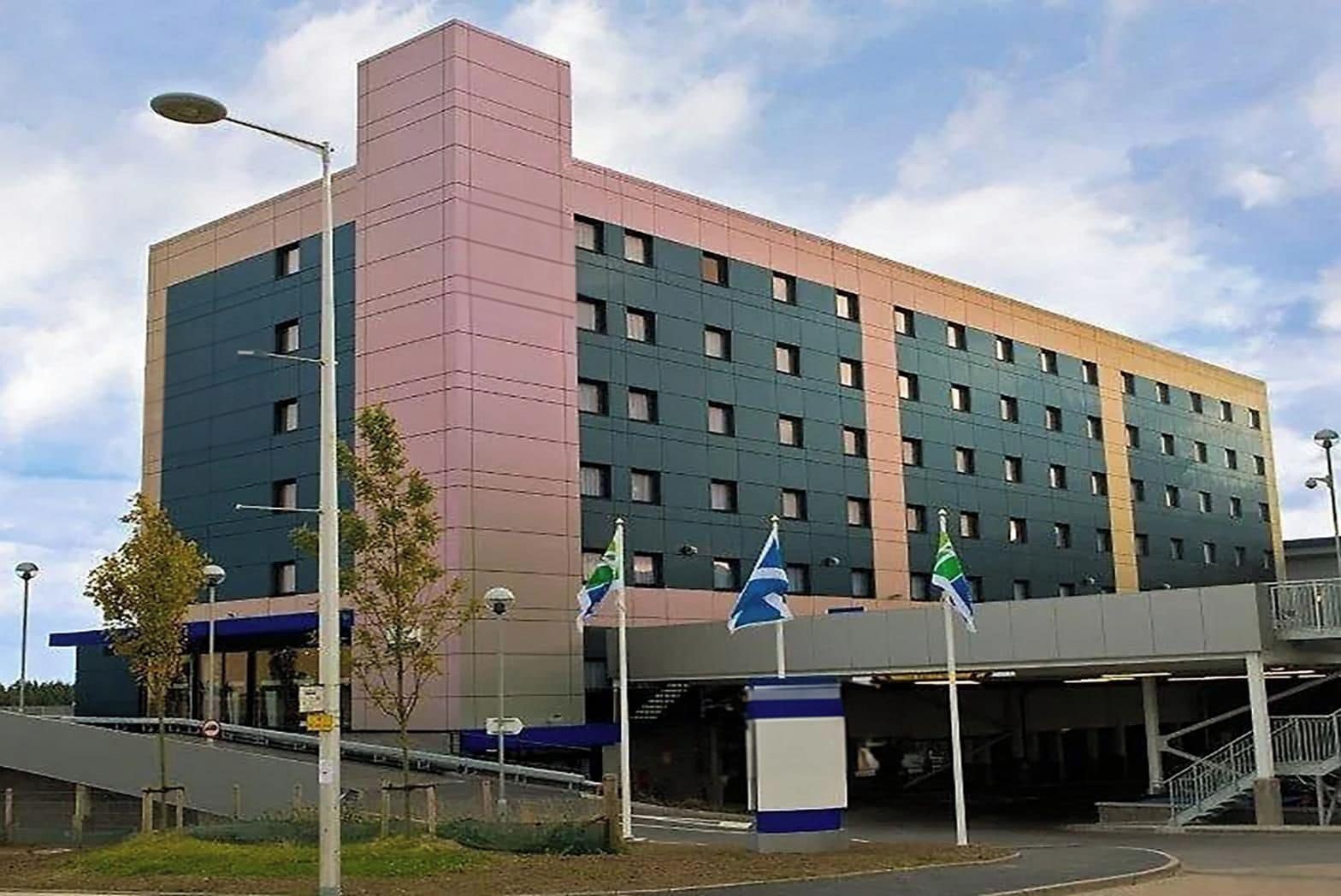 A multi-story hotel with a modern design, featuring a prominent pink and green facade, multiple windows, and a covered entrance area with flags.