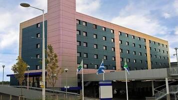 A multi-story hotel with a modern design, featuring a prominent pink and green facade, multiple windows, and a covered entrance area with flags.