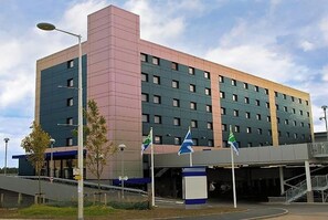 A multi-story hotel with a modern design, featuring a prominent pink and green facade, multiple windows, and a covered entrance area with flags.