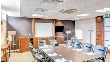 A conference room with a long table, multiple chairs, a projector screen, a TV, and a whiteboard.