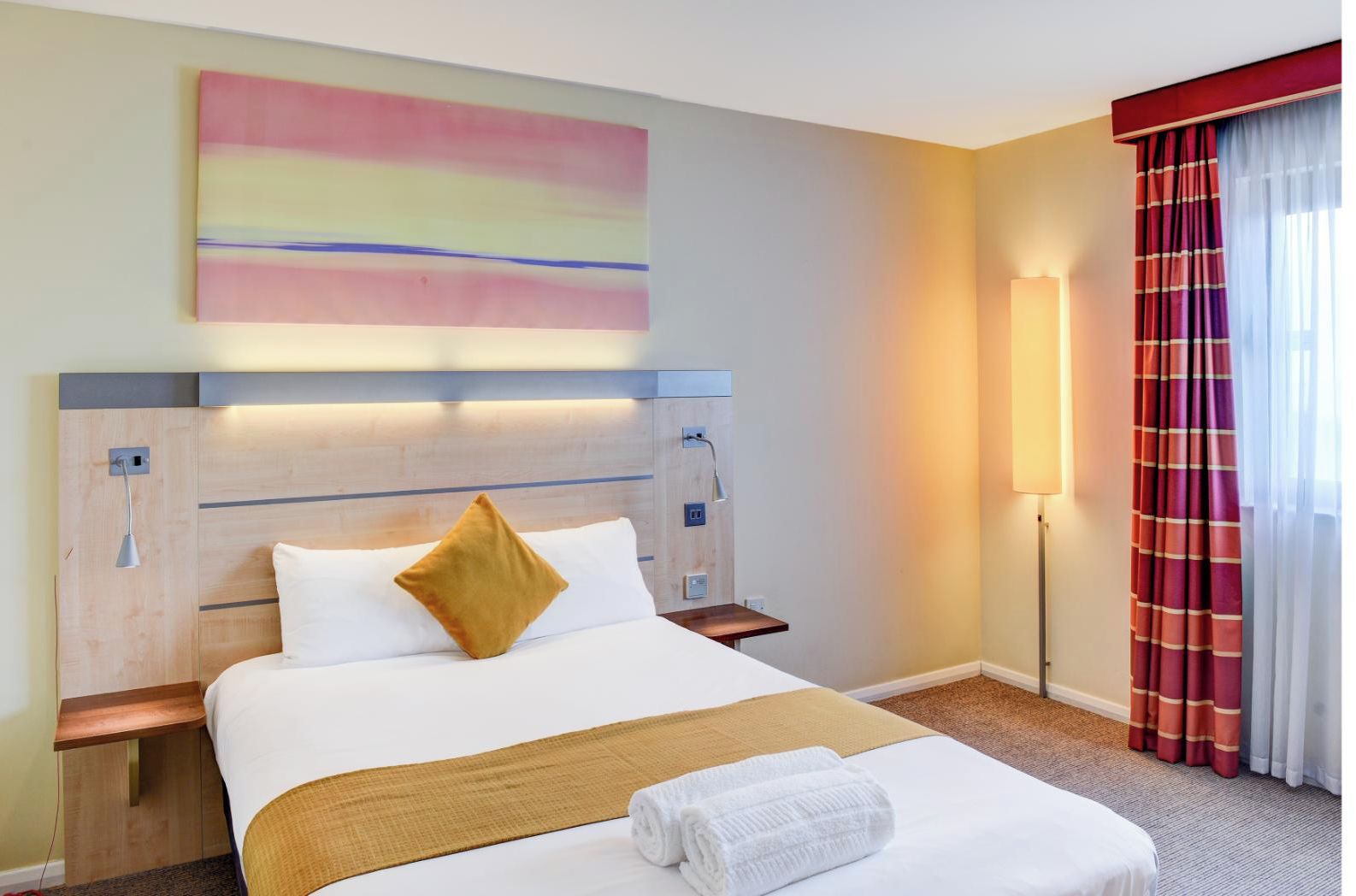 A hotel room with a large bed, a headboard with built-in lighting, a bedside table, a wall-mounted lamp, and a window with curtains.
