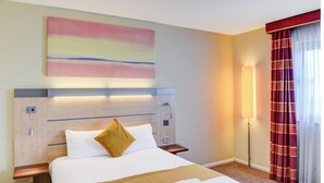 Une chambre d’hôtel avec un grand lit, une tête de lit dotée d’un éclairage intégré, une table de chevet, une lampe fixée au mur et une fenêtre avec des rideaux.