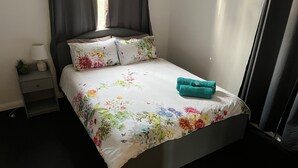 Una cama bien tendida con ropa de cama floral, una toalla color turquesa y una mesita de noche con una lámpara y una planta pequeña.