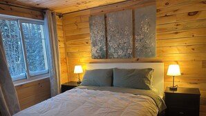 A cozy wooden room with a bed, two bedside tables with lamps, a window with curtains, and a painting on the wall.