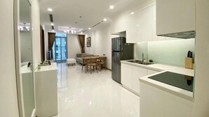 A modern kitchen with white cabinets, a stainless steel refrigerator, and a dining area with a table and chairs. - Vinhomes Central Park AnAn Apartment (Ho Chi Minh City)