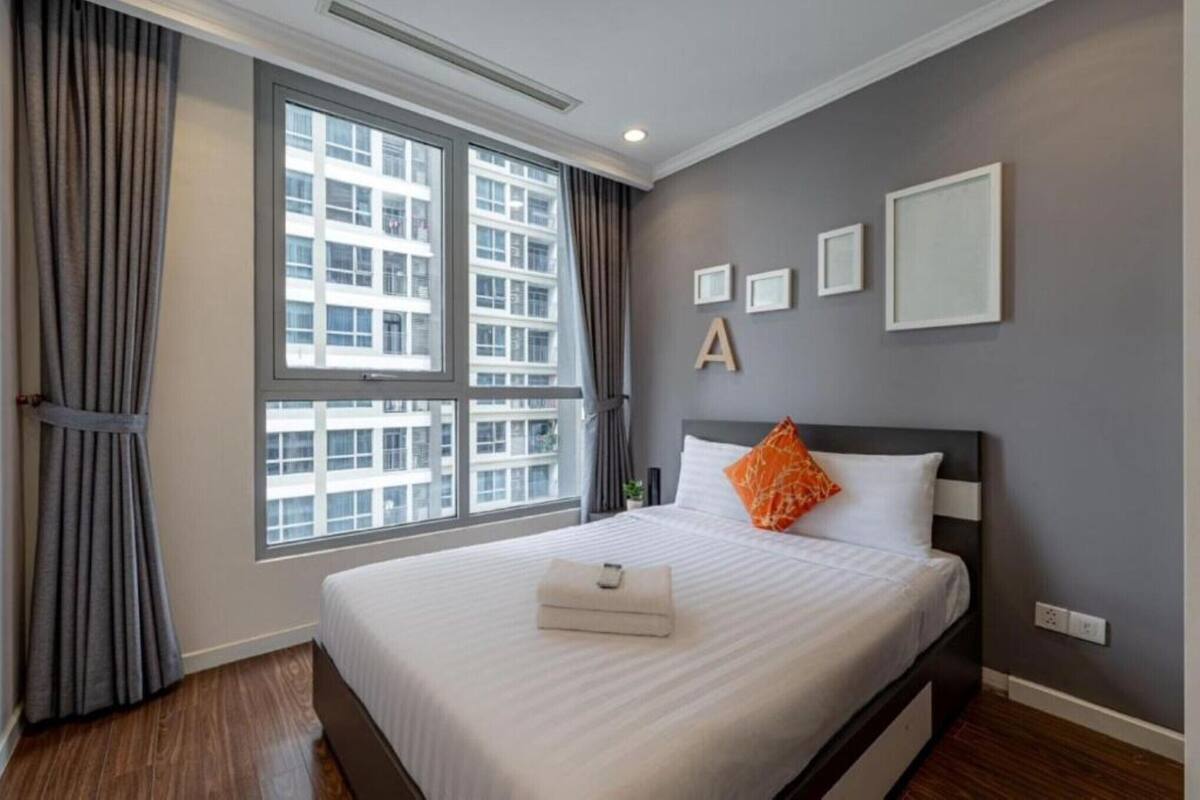 Una habitación de hotel con un ventanal grande, una cama con ropa blanca y una almohada naranja, y una pared gris con tres cuadros enmarcados.