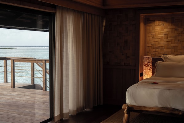 Un dormitorio con piso de madera, una cama con sábanas blancas, vista al océano por una ventana y una pared de madera con diseño tejido.