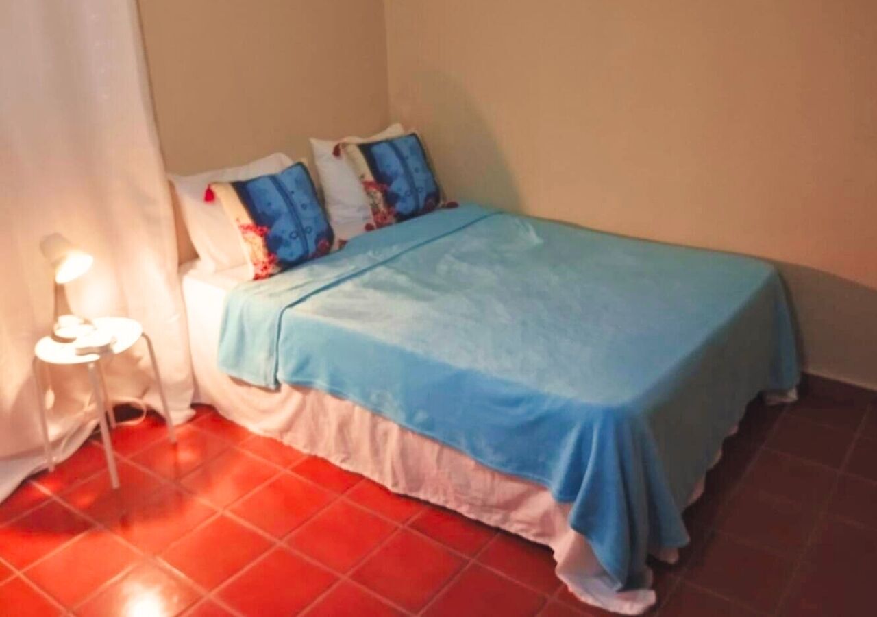 Una habitación pequeña con cama individual, colcha azul, almohadas blancas y piso de baldosas rojas.