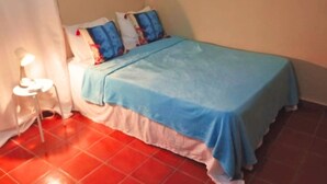 Une petite chambre avec un lit simple, une couvre-lit bleue, des oreillers blancs et un sol carrelé de carreaux rouges.