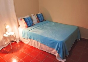 Een kleine kamer met een enkel bed, een blauwe spreidek, witte kussens en een vloer van rode tegels.