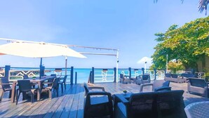 Une terrasse en bois avec des espaces de détente en plein air, comprenant des tables et des chaises, donnant sur une étendue d’eau.