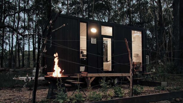 Una cabaña pequeña con un hoyo para fuego, terraza de madera y una puerta que da a un espacio interior.