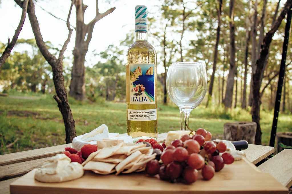 Uma montagem para piquenique com uma garrafa de vinho italiano, um copo, e diversos queijos e uvas sobre uma mesa de madeira em uma área arborizada.