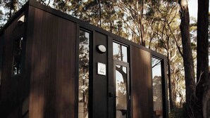 Una cabaña pequeña y oscura de madera, con ventanas grandes, una terraza de madera y una puerta.