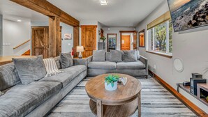 A modern living room with a sectional sofa, a round wooden coffee table, and a large window with a view of a rocky shoreline. - Kings Beach Retreat | Hot Tub & Game Loft (Kings Beach)