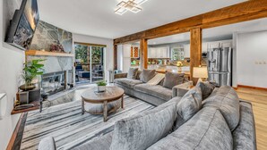 A spacious living room with a large gray sectional sofa, a round coffee table, and a mounted flat-screen TV. The room features a wooden ceiling beam, a stone fireplace, and a large window offering a view of the outdoors. - Kings Beach Retreat | Hot Tub & Game Loft (Kings Beach)