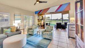 A spacious living area with a sofa, armchairs, a coffee table, and a ceiling fan. A large window offers a view of the outdoors.