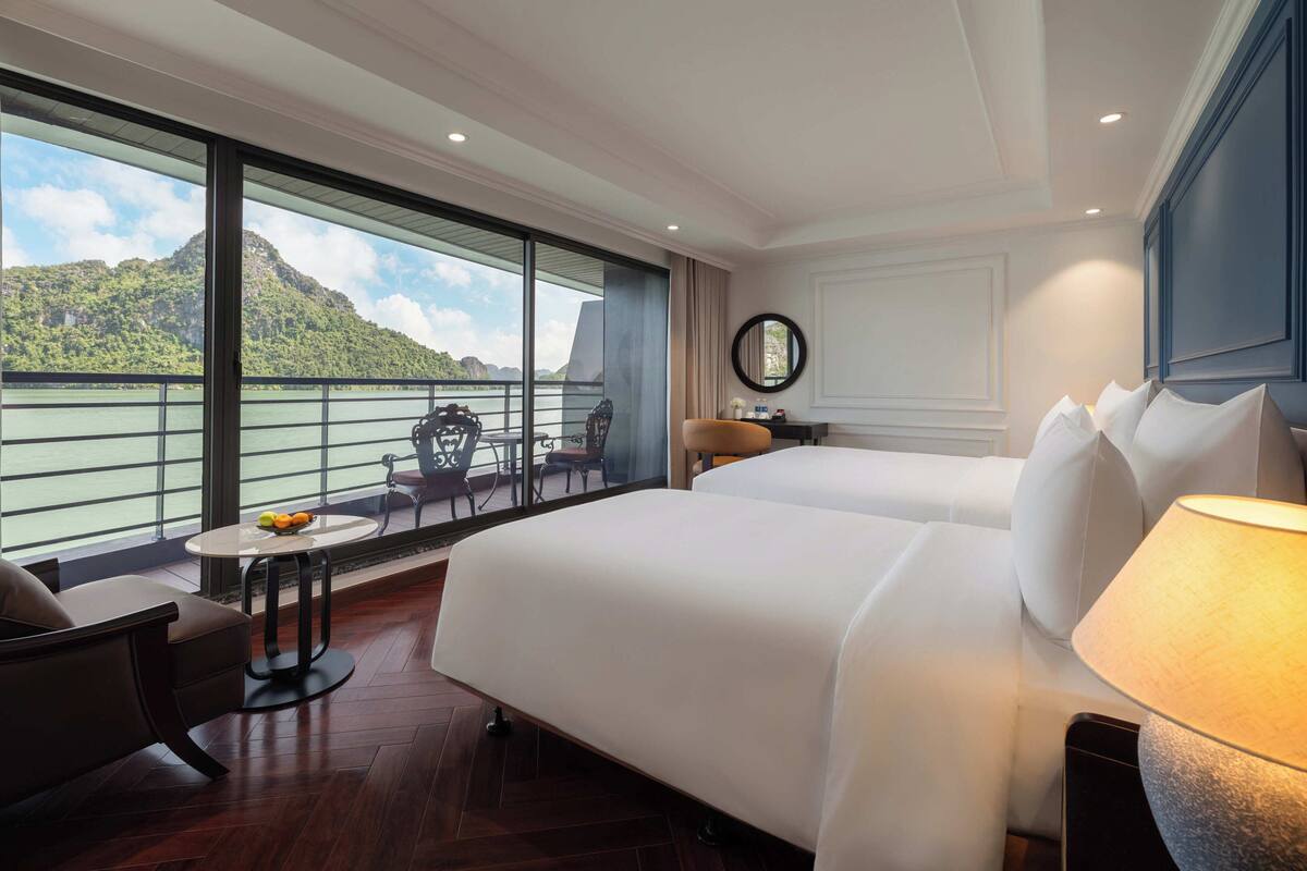 A hotel room with two beds, a small table, a chair, a lamp, and a large window offering a view of a body of water and mountains.