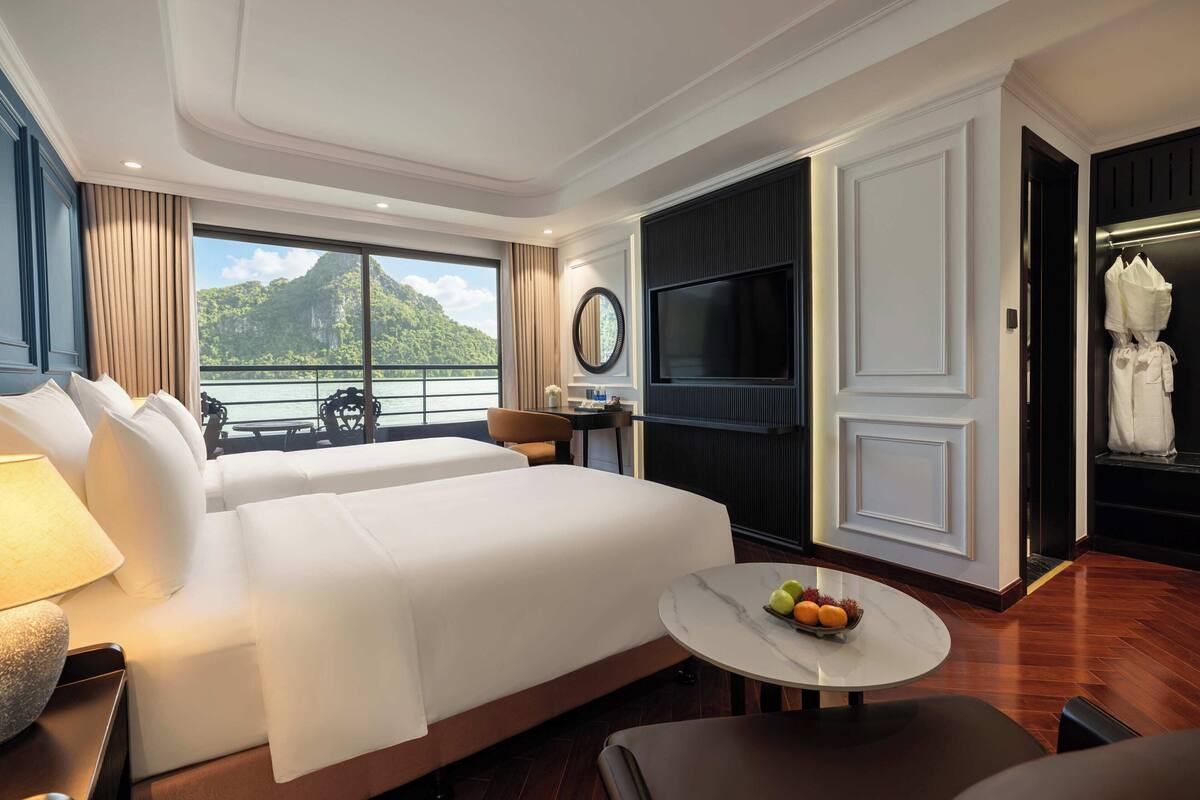 A hotel room with a bed, a desk, a TV, a round table with fruit, and a view of the sea.