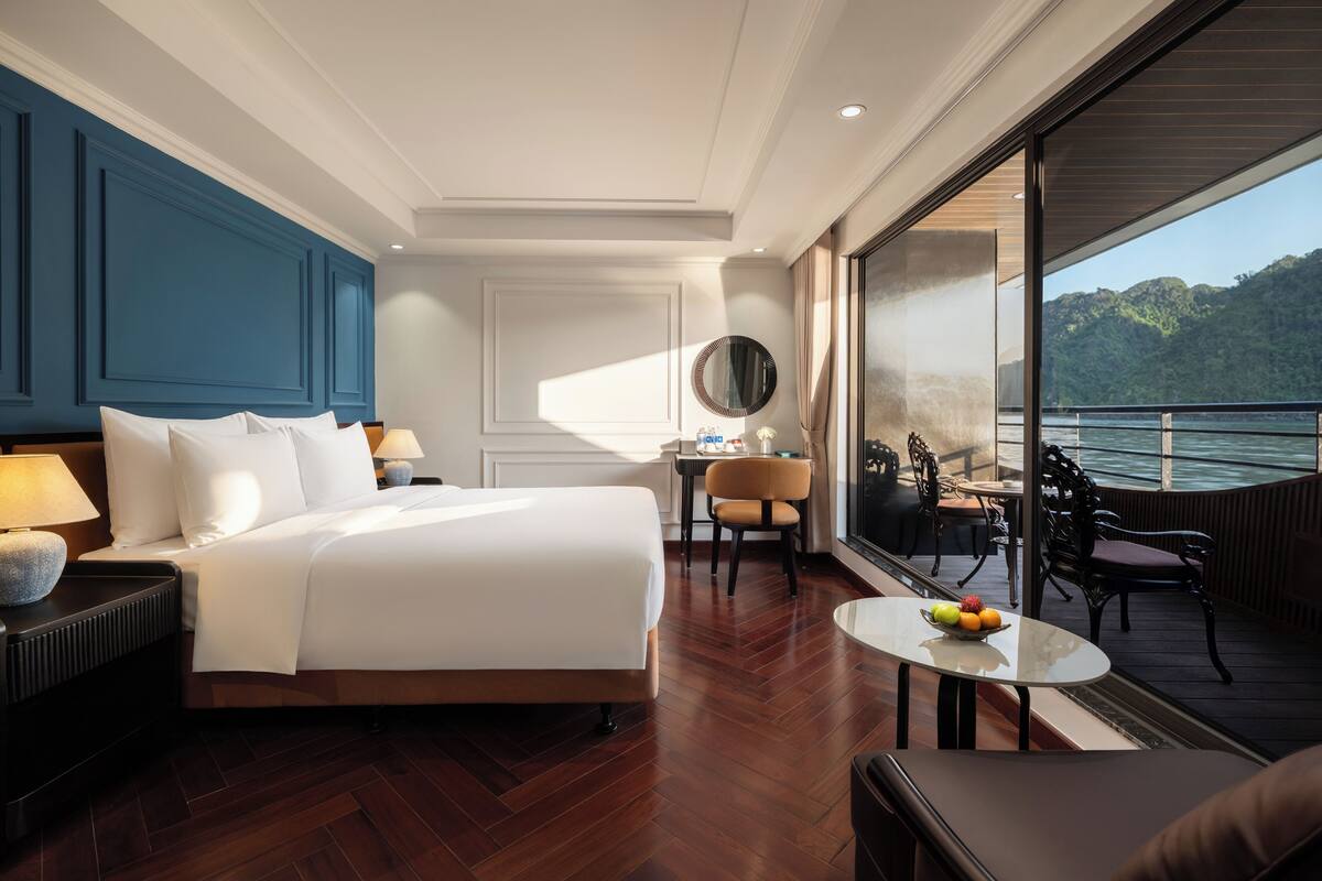 A hotel room with a large bed, a desk, a chair, a small table with fruit, and a balcony with a view of the water.