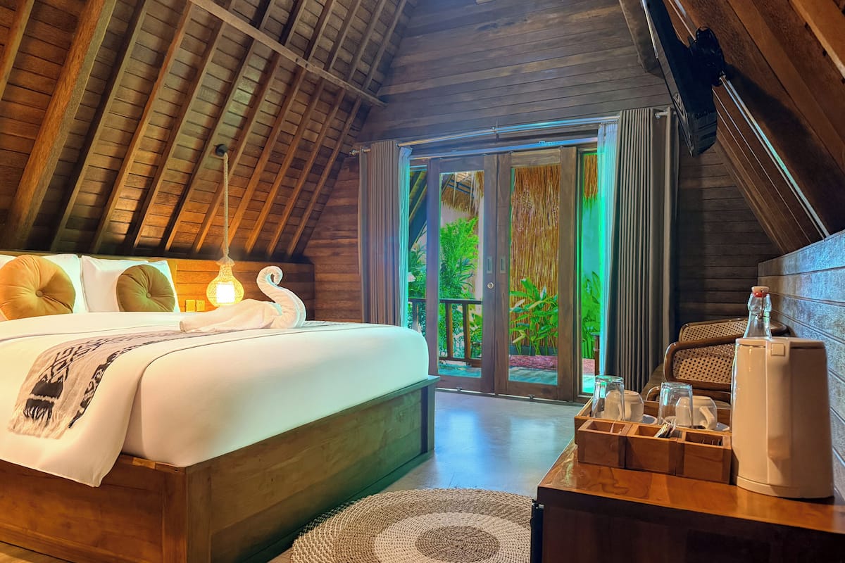 A bedroom with a wooden ceiling, a bed with white bedding, a bedside table with a lamp, and a view of greenery outside.