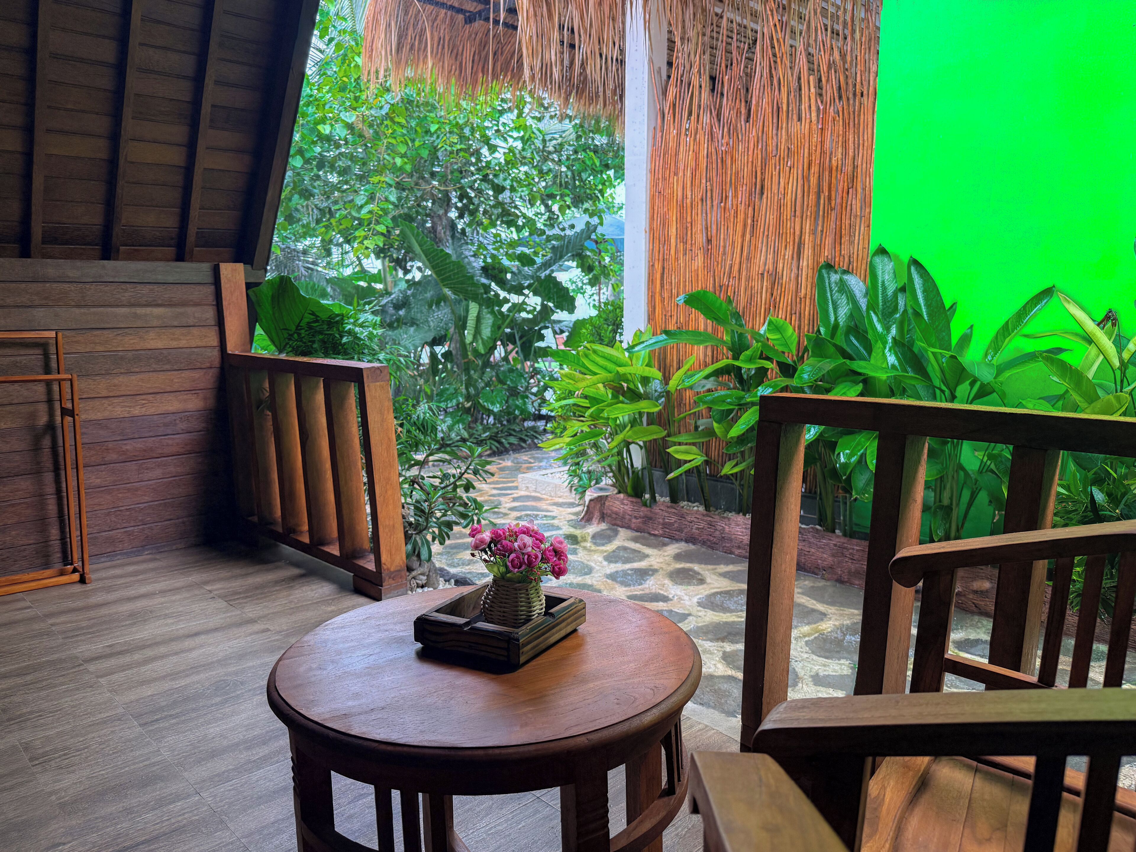 A wooden table with a vase of flowers, a wooden bench, and a view of a lush garden with a bamboo fence and tropical plants.