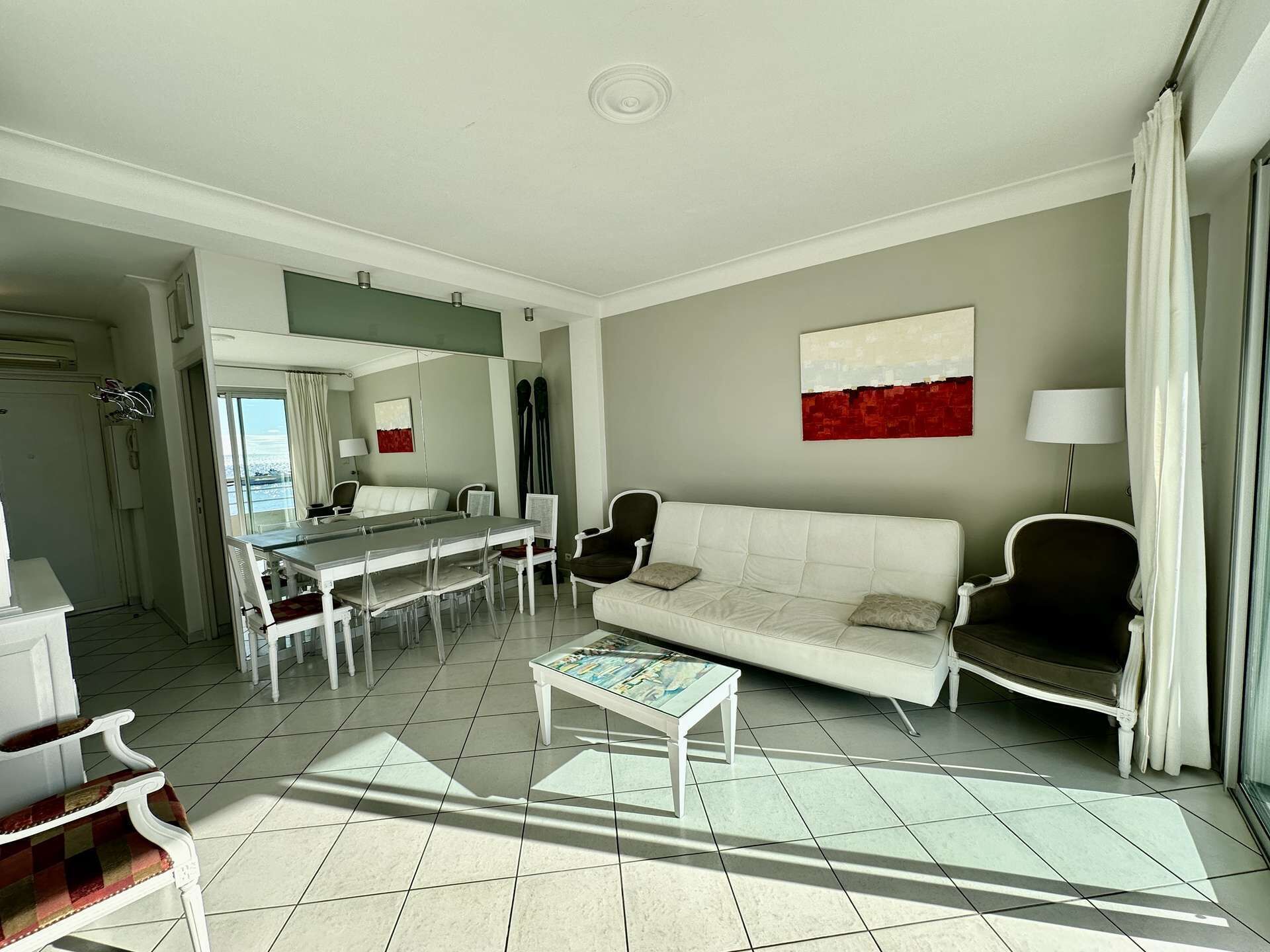 A modern living room with a white sofa, a dining table with chairs, a coffee table, and a large window with curtains.