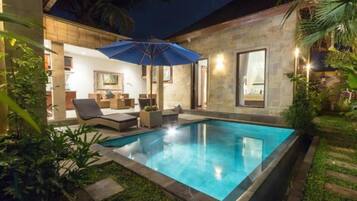 A swimming pool with lounge chairs and an umbrella, surrounded by greenery and a stone building.