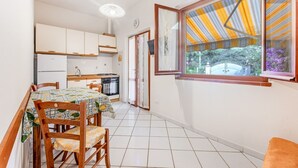 A compact kitchen with white cabinets, a dining table with chairs, and a window with a striped awning. - Casa dei Fiori - Goelba (Portoferraio)